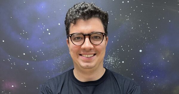 Uma pessoa com o rosto desfocado está em pé diante de um fundo que lembra o espaço sideral, com estrelas e nebulosas visíveis. A pessoa veste uma camiseta preta. (varejo)