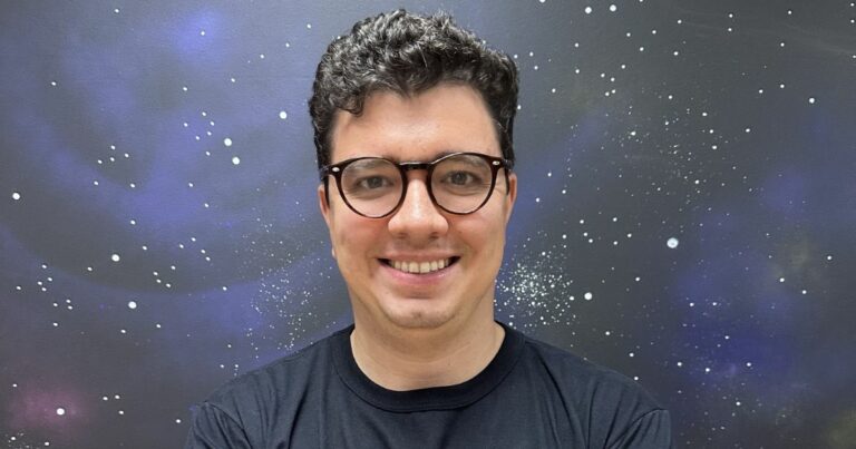 Uma pessoa com o rosto desfocado está em pé diante de um fundo que lembra o espaço sideral, com estrelas e nebulosas visíveis. A pessoa veste uma camiseta preta. (varejo)