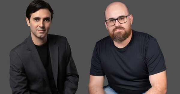 Duas pessoas estão sentadas lado a lado diante de um fundo cinza neutro. A pessoa à esquerda veste um terno preto com uma camiseta preta por baixo, transmitindo uma aparência mais formal. Já a pessoa à direita opta por um estilo casual, com uma camiseta preta de mangas curtas e calça jeans. (Dynadok)