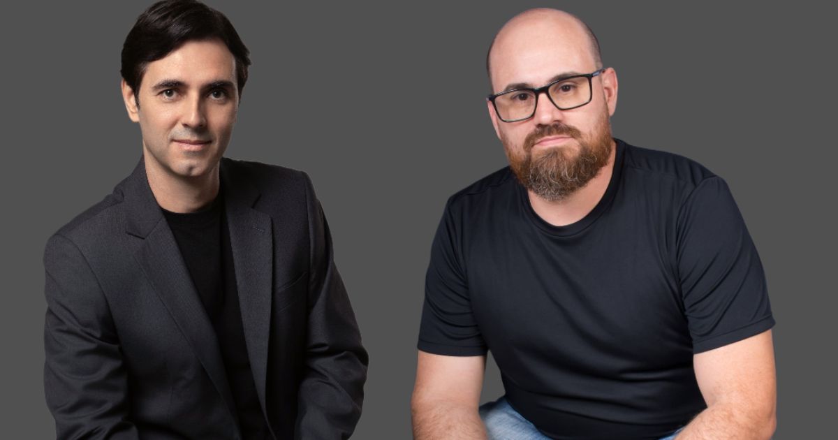 Duas pessoas estão sentadas lado a lado diante de um fundo cinza neutro. A pessoa à esquerda veste um terno preto com uma camiseta preta por baixo, transmitindo uma aparência mais formal. Já a pessoa à direita opta por um estilo casual, com uma camiseta preta de mangas curtas e calça jeans. (Dynadok)
