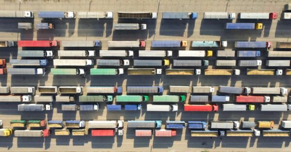 Imagem aérea de uma ampla estrada pavimentada com múltiplas faixas, onde diversos caminhões estão alinhados em filas paralelas. Os veículos variam em cor e tamanho, e estão organizados de forma ordenada, sugerindo uma área de espera ou estacionamento. A luz do sol projeta sombras nítidas dos caminhões sobre o asfalto, indicando que a foto foi tirada durante o dia. Não há presença de pessoas, textos ou outros elementos visuais além dos caminhões e da estrada. (Rumo e Nstech)