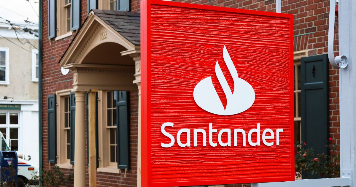 A imagem mostra uma placa vermelha com o logotipo do banco Santander e a palavra "Santander" escrita em branco. Ao fundo, há um prédio de tijolos com janelas e uma porta visível, além de outro edifício ao longe.