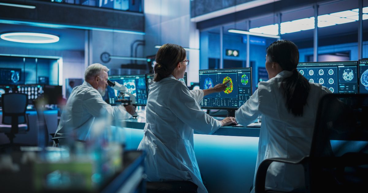 Três pessoas usando jalecos brancos estão sentadas em frente a uma mesa com vários monitores de computador em um ambiente de laboratório moderno. As telas exibem imagens científicas, como exames cerebrais e outros dados médicos. Ao fundo, é possível ver equipamentos de laboratório, reforçando o cenário tecnológico e voltado à pesquisa científica. (IGEN)