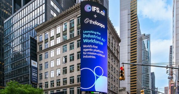 Um grande painel digital está instalado na lateral de um prédio em uma área urbana. O painel exibe um anúncio das empresas IFS.ai e theloops, anunciando o lançamento da "Industrial AI Workforce". O texto no painel diz: "IFS.ai + theloops Launching the Industrial AI Workforce World's first agentic platform for mission-critical industries."