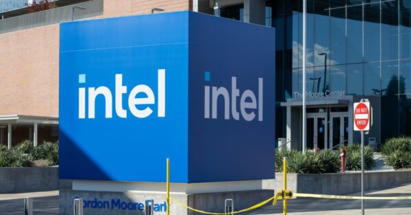 Um grande cubo azul com o logotipo branco da Intel está posicionado em frente a um edifício moderno com fachada de vidro. O cubo está em uma área externa, cercado por vegetação e delimitado por uma fita de segurança amarela. Na base do cubo, lê-se "Gordon Moore Park". Ao fundo, a entrada do prédio exibe o nome "The Moore Center", e há uma placa de trânsito com os dizeres "DO NOT ENTER".