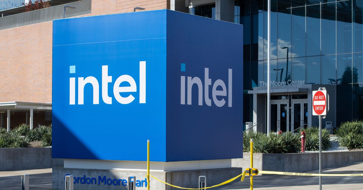 Um grande cubo azul com o logotipo branco da Intel está posicionado em frente a um edifício moderno com fachada de vidro. O cubo está em uma área externa, cercado por vegetação e delimitado por uma fita de segurança amarela. Na base do cubo, lê-se "Gordon Moore Park". Ao fundo, a entrada do prédio exibe o nome "The Moore Center", e há uma placa de trânsito com os dizeres "DO NOT ENTER".