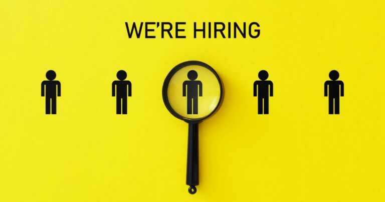 Imagem com fundo amarelo vibrante. No centro, há cinco ícones de figuras humanas na cor preta, dispostos lado a lado em linha horizontal. Acima delas, em letras maiúsculas pretas, está a frase "WE'RE HIRING" (Estamos contratando). Uma lupa preta está posicionada sobre a figura central, destacando-a como se estivesse sendo selecionada ou analisada. (vagas)