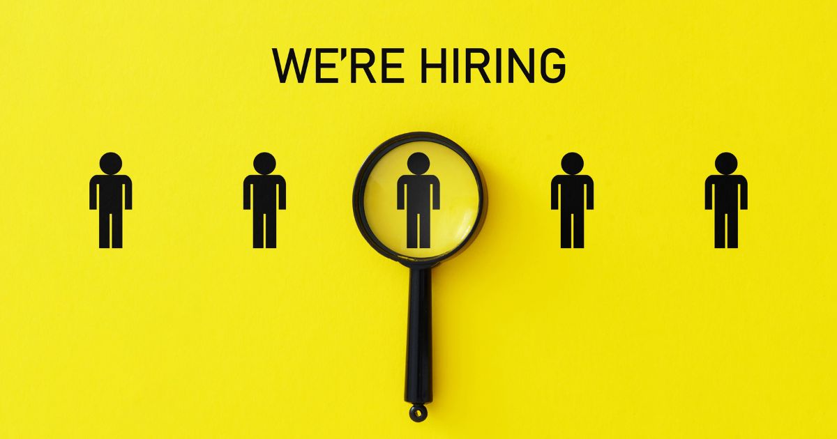 Imagem com fundo amarelo vibrante. No centro, há cinco ícones de figuras humanas na cor preta, dispostos lado a lado em linha horizontal. Acima delas, em letras maiúsculas pretas, está a frase "WE'RE HIRING" (Estamos contratando). Uma lupa preta está posicionada sobre a figura central, destacando-a como se estivesse sendo selecionada ou analisada. (vagas)