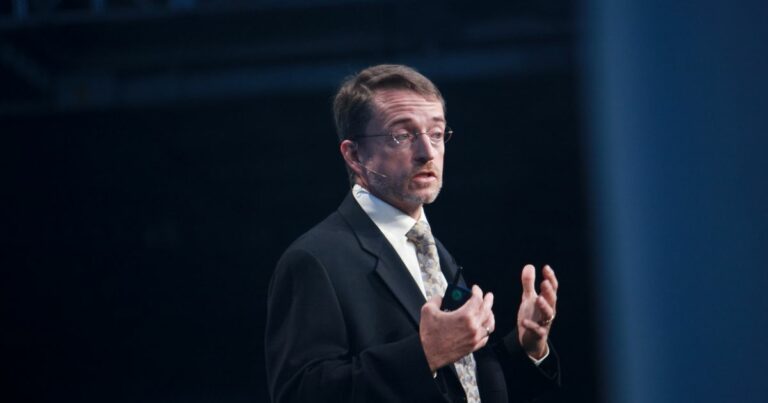 Pat Gelsinger, ex-CEO da Intel, vestido com terno e gravata está em pé, gesticulando com ambas as mãos. O rosto da pessoa está desfocado. O fundo é escuro, sugerindo um ambiente interno, possivelmente durante uma apresentação ou discurso. (Intel)