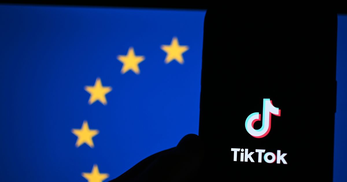 Silhueta de um smartphone exibindo o logotipo do TikTok, posicionada em frente a um fundo com a bandeira da União Europeia — um campo azul com um círculo formado por doze estrelas amarelas.