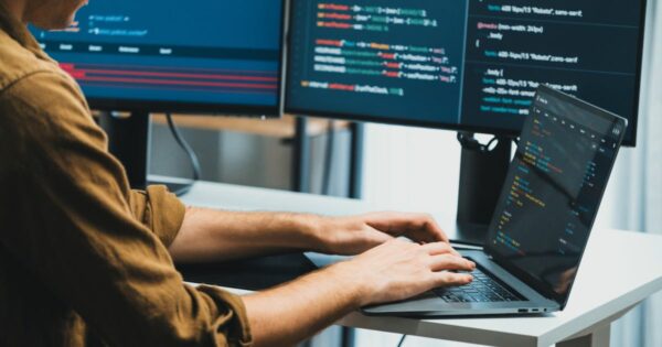 2025, Uma pessoa está trabalhando em um laptop conectado a dois monitores externos, que exibem linhas de código em diferentes cores. O desenvolvedor digita no teclado do laptop, e os três monitores mostram trechos de programação. O ambiente sugere um espaço de trabalho voltado para desenvolvimento de software (brasil, brasscom, treinee, digitização)