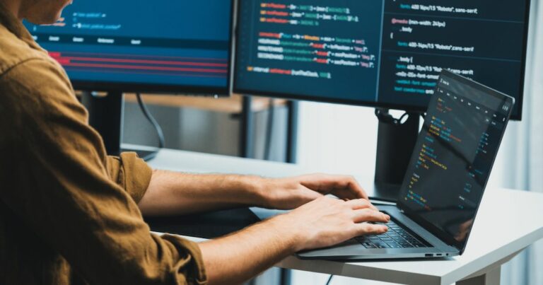 2025, Uma pessoa está trabalhando em um laptop conectado a dois monitores externos, que exibem linhas de código em diferentes cores. O desenvolvedor digita no teclado do laptop, e os três monitores mostram trechos de programação. O ambiente sugere um espaço de trabalho voltado para desenvolvimento de software (brasil, brasscom, treinee, digitização)