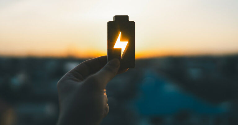 Imagem de uma mão segurando um objeto em forma de bateria com um símbolo de raio recortado no centro, através do qual a luz do pôr do sol brilha. Ao fundo, a paisagem urbana aparece desfocada. A imagem transmite uma ideia de energia ou recarga associada à luz natural (energia, bioenergia, brasil, EDP)
