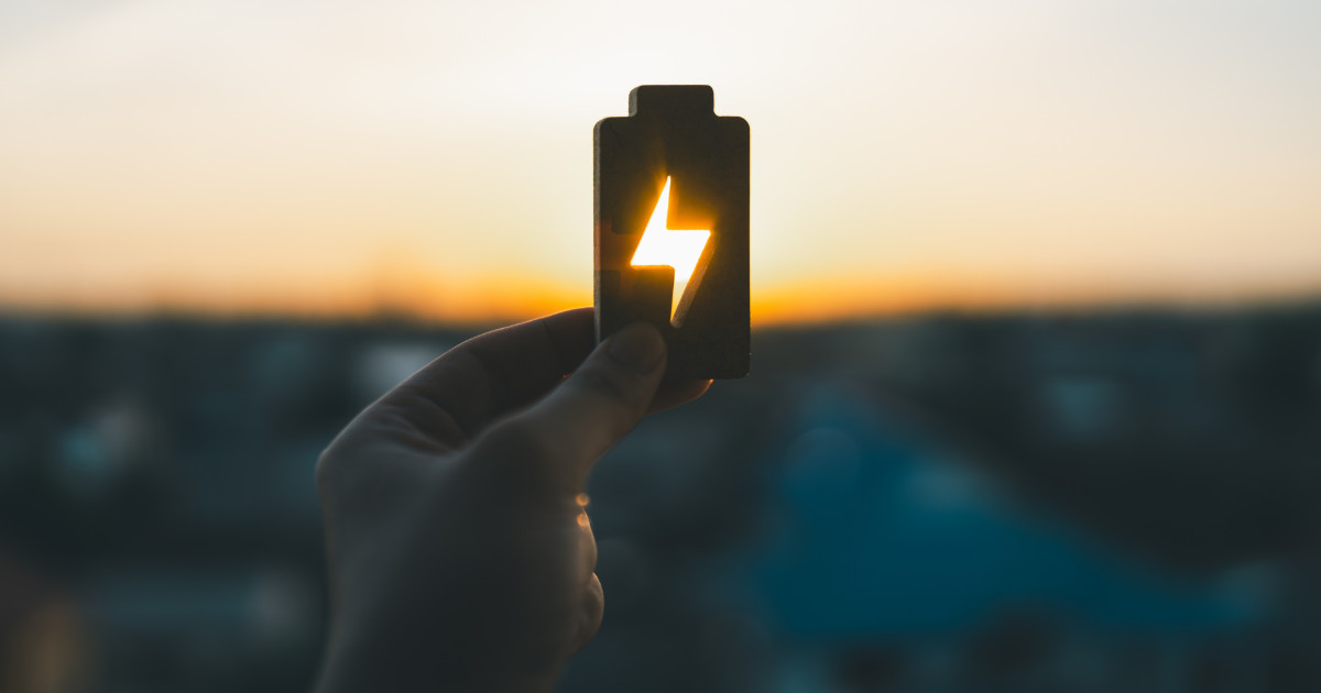 Imagem de uma mão segurando um objeto em forma de bateria com um símbolo de raio recortado no centro, através do qual a luz do pôr do sol brilha. Ao fundo, a paisagem urbana aparece desfocada. A imagem transmite uma ideia de energia ou recarga associada à luz natural (energia, bioenergia, brasil, EDP)