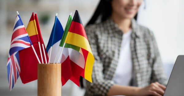 Foto mostra pequenas bandeiras de diversos países — incluindo Reino Unido, Espanha, França, Itália e Alemanha — em um suporte de madeira, com uma mulher sorridente ao fundo usando fone de ouvido e trabalhando em um laptop, representando aprendizado de idiomas.