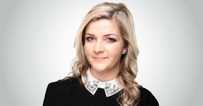 Na imagem, Patrycja Sobera, Vice-presidente Global de Digital Workplace Solutions da Unisys. Ela tem cabelo loiro ondulado, usando uma blusa preta com gola branca adornada com pedras brilhantes. (presencial)