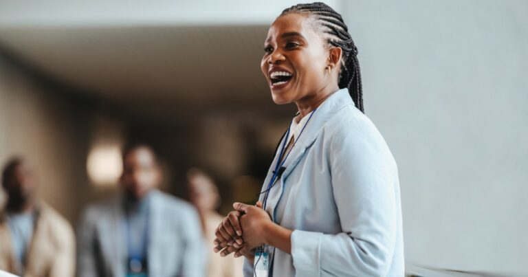 Mulher sorridente de perfil, usando jaleco claro e crachá, aparentemente em ambiente corporativo ou conferência. Ela está falando com um pequeno grupo de pessoas desfocadas ao fundo, transmitindo confiança e entusiasmo (empresária, profissional)