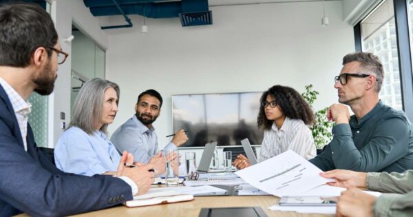 Imagem de uma reunião estratégica em uma sala de conferências moderna, com seis pessoas de diferentes idades e etnias sentadas ao redor de uma mesa. Todos estão envolvidos em uma discussão, com papéis, notebooks e copos de água sobre a mesa. Ao fundo, há uma grande tela de TV desligada e janelas que mostram prédios externos. O ambiente transmite profissionalismo e colaboração (estratégia, segurança)