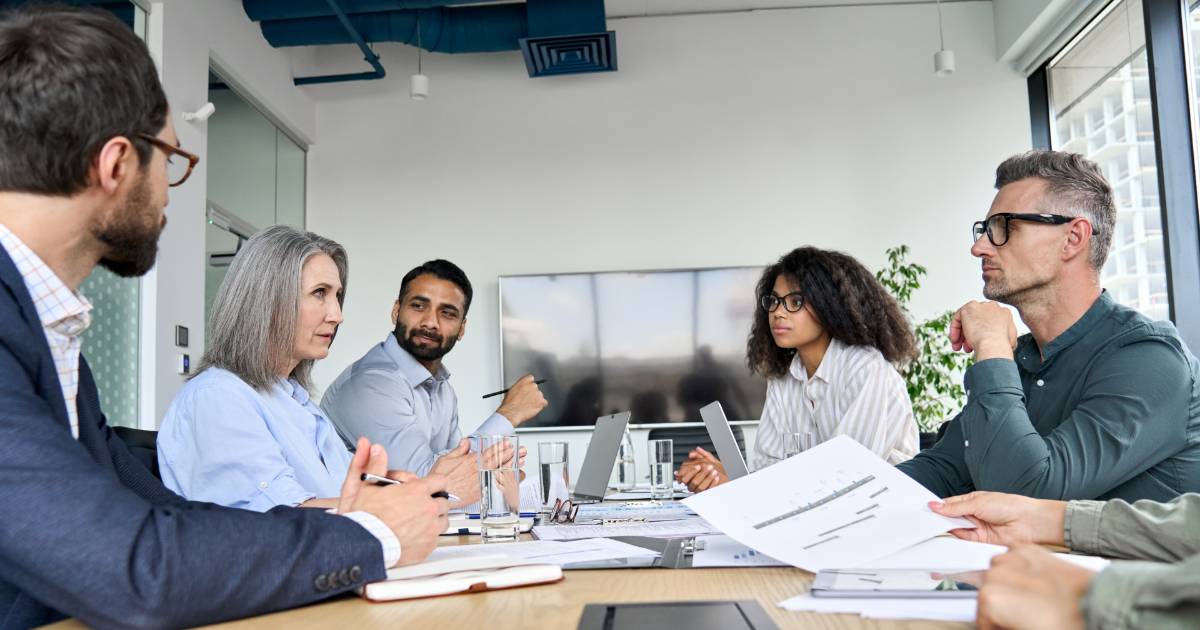 Imagem de uma reunião estratégica em uma sala de conferências moderna, com seis pessoas de diferentes idades e etnias sentadas ao redor de uma mesa. Todos estão envolvidos em uma discussão, com papéis, notebooks e copos de água sobre a mesa. Ao fundo, há uma grande tela de TV desligada e janelas que mostram prédios externos. O ambiente transmite profissionalismo e colaboração (estratégia, segurança)