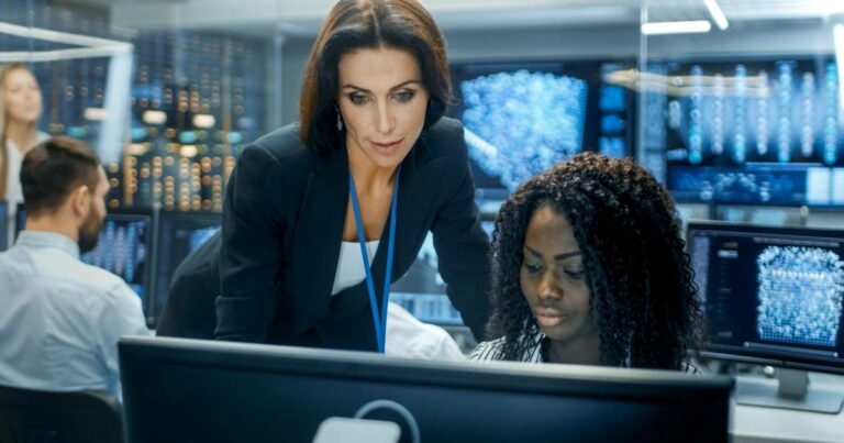 Duas mulheres trabalhando em frente a um computador em um centro de operações de segurança digital; uma delas, identificada como líder de segurança (CSO), está de pé e orientando a outra, que está sentada. Ao fundo, há vários monitores exibindo dados e gráficos digitais, sugerindo um ambiente de alta tecnologia voltado à cibersegurança (prevenção, dados, infraestrutura)