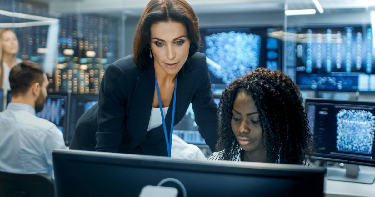 Duas mulheres trabalhando em frente a um computador em um centro de operações de segurança digital; uma delas, identificada como líder de segurança (CSO), está de pé e orientando a outra, que está sentada. Ao fundo, há vários monitores exibindo dados e gráficos digitais, sugerindo um ambiente de alta tecnologia voltado à cibersegurança (prevenção, dados, infraestrutura)