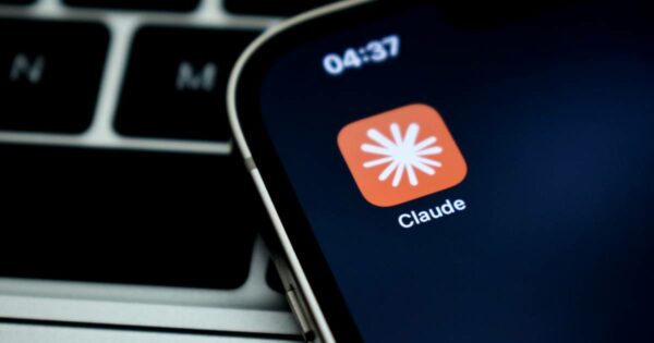 Ícone do aplicativo Claude, da empresa Anthropic, exibido na tela de um smartphone. O ícone é laranja com um símbolo branco estilizado no centro, lembrando um asterisco ou explosão, com o nome "Claude" escrito abaixo. Ao fundo, vê-se parcialmente o teclado de um laptop. IBM Mythos