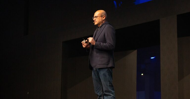 Cleber Morais, diretor-geral da AWS no Brasil, em pé no palco durante uma apresentação, vestindo blazer azul-escuro e calça jeans, usando microfone de lapela e segurando um controle remoto, com fundo escuro e iluminação pontual (maioria)