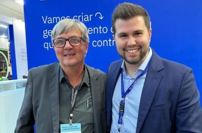 Duas pessoas estão posicionadas lado a lado, ambas vestindo trajes sociais. Ao fundo, há uma parede azul com texto em português que diz: "Vamos criar o gerenciamento de qualidade que você controla." Não há elementos matemáticos ou escolares visíveis na imagem. (pais)