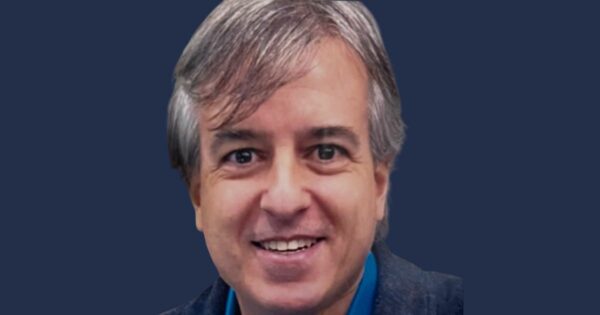 Retrato de Ricardo Ribeiro, CIO da Movecta, sorrindo. Ele está com uma camisa azul e blazer escuro, posando diante de um fundo azul sólido.