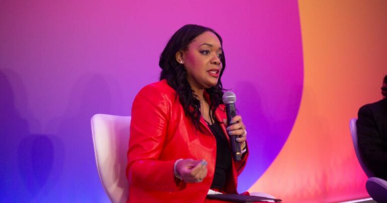 Francessca Vasquez, vice-presidente de Serviços Profissionais e Agentes de IA da Amazon Web Services (AWS), falando ao microfone durante um evento, vestindo blazer vermelho e blusa preta, sentada em cadeira branca com fundo em tons de roxo, rosa e laranja.