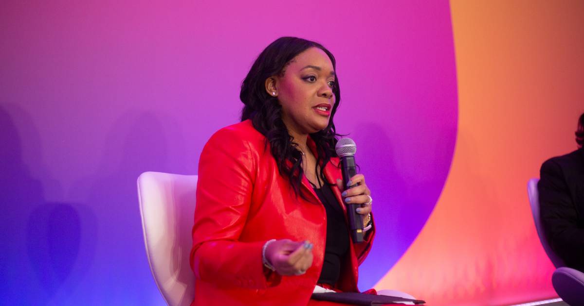 Francessca Vasquez, vice-presidente de Serviços Profissionais e Agentes de IA da Amazon Web Services (AWS), falando ao microfone durante um evento, vestindo blazer vermelho e blusa preta, sentada em cadeira branca com fundo em tons de roxo, rosa e laranja.