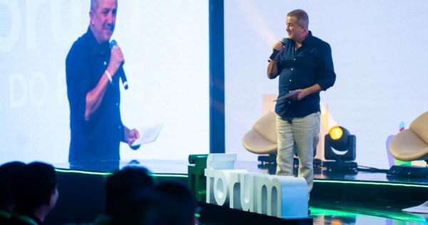 Adelson de Sousa, fundador e presidente do Itaqui, fala ao microfone no palco durante a abertura do IT Forum Praia do Forte, segurando um papel nas mãos, com o logotipo do evento em destaque à frente do palco.