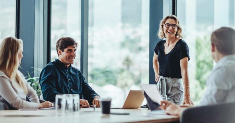 Imagem de uma reunião de trabalho em um escritório moderno, com quatro pessoas interagindo de forma descontraída. Uma mulher em pé, sorridente e confiante, conversa com os colegas, transmitindo energia positiva e liderança, enquanto os outros participantes, sentados à mesa com notebooks e documentos, também sorriem e participam da conversa (humano, empresas)
