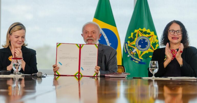 O presidente Luiz Inácio Lula da Silva aparece ao centro segurando um documento aberto durante a sanção do Projeto de Lei nº 847/2025, que aprimora a destinação de recursos do Fundo Nacional de Desenvolvimento Científico e Tecnológico (FNDCT). Ao lado esquerdo da imagem está a ministra Gleisi Hoffmann e, à direita, a ministra Luciana Santos, ambas aplaudindo. Ao fundo, estão as bandeiras do Brasil e da República. A cena ocorre em uma sala de reuniões com mesa de madeira refletindo os participantes. (Projetos)