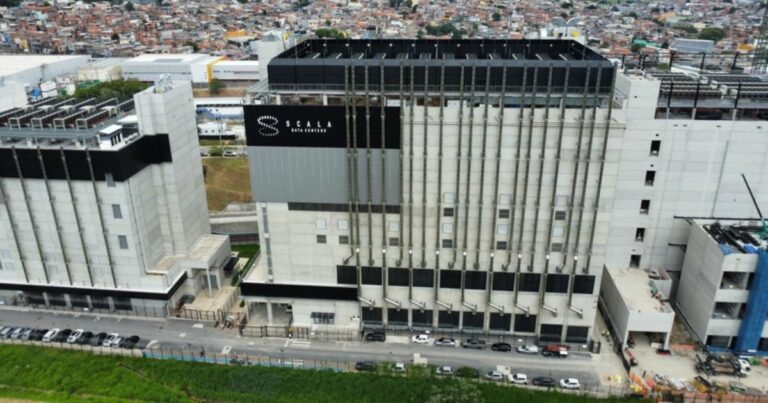 A imagem mostra um grande complexo industrial ou comercial composto por várias estruturas. O edifício principal, localizado ao centro, possui uma placa com os dizeres "SCALA DATA CENTERS". Há diversos carros estacionados ao longo da rua em frente aos prédios, e ao fundo é possível ver uma área urbana densamente povoada.