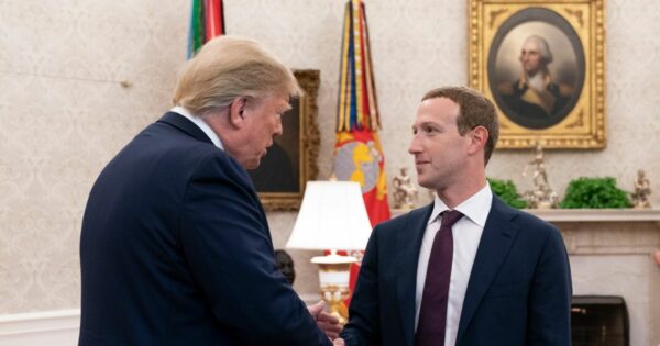 Dois homens vestindo ternos estão apertando as mãos em um ambiente formal, possivelmente um escritório ou sala de reuniões. Ao fundo, há uma luminária, bandeiras e um retrato emoldurado pendurado na parede. (Zuckerberg)