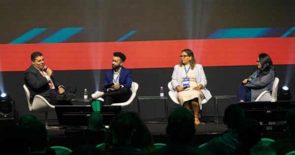 Quatro pessoas estão sentadas em um palco, participando de uma discussão em painel ou entrevista. Ao fundo, há uma tela grande com gráficos abstratos em azul e vermelho. (cibersegurança)