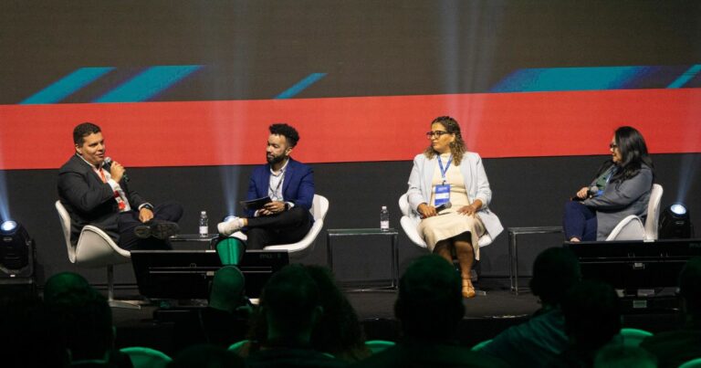 Quatro pessoas estão sentadas em um palco, participando de uma discussão em painel ou entrevista. Ao fundo, há uma tela grande com gráficos abstratos em azul e vermelho. (cibersegurança)