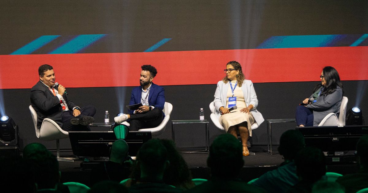 Quatro pessoas estão sentadas em um palco, participando de uma discussão em painel ou entrevista. Ao fundo, há uma tela grande com gráficos abstratos em azul e vermelho. (cibersegurança)