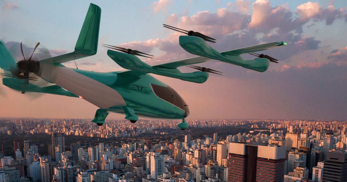 Uma aeronave elétrica de decolagem e pouso vertical (eVTOL), na cor verde-azulada, está voando sobre uma cidade densamente povoada, com muitos prédios altos. O céu está parcialmente nublado, com tons quentes de laranja e rosa, sugerindo o nascer ou o pôr do sol. (Eve)