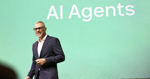 Uma pessoa está em pé diante de um grande painel verde que exibe o texto "AI Agents" em letras brancas. Ela veste um terno escuro com uma camisa branca, transmitindo uma aparência formal e profissional. (Zendesk)