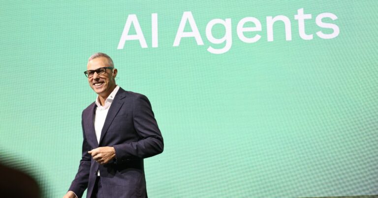 Uma pessoa está em pé diante de um grande painel verde que exibe o texto "AI Agents" em letras brancas. Ela veste um terno escuro com uma camisa branca, transmitindo uma aparência formal e profissional. (Zendesk)