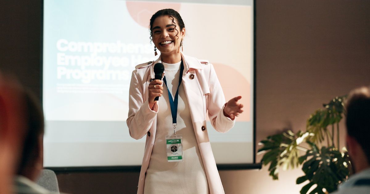 Uma pessoa está em pé diante de uma tela, segurando um microfone, aparentemente conduzindo uma apresentação. Na tela ao fundo, lê-se o título "Comprehensive Employee Training Program". A pessoa veste uma roupa branca com um casaco rosa claro e usa um crachá pendurado no pescoço. Na frente, há figuras desfocadas que indicam a presença de uma plateia. À direita da imagem, é possível ver uma planta decorativa (vagas)