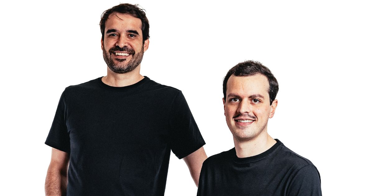 Duas pessoas estão presentes na imagem, ambas vestindo camisetas pretas. A pessoa à esquerda está em pé, enquanto a pessoa à direita está sentada. O fundo é completamente branco, sem elementos adicionais visíveis. (Voa Health)