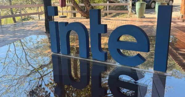 Letreiro tridimensional azul com a palavra "intel" sobre uma superfície refletiva, exibindo o reflexo das letras. Ao fundo, há um deque de madeira, árvores e um cenário externo ensolarado. (parceiro, Intel)