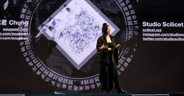 Sougwen Chung, artista e pesquisadora sino-canadense, apresenta sua palestra no palco do IT Forum Praia do Forte 2025. Ela está em pé, gesticulando com as mãos enquanto fala, vestindo blazer escuro sobre blusa preta. Ao fundo, telas exibem padrões visuais em tons de azul que remetem a ondas ou fluxos digitais, criando uma atmosfera tecnológica que complementa sua apresentação sobre arte e inteligência artificial.