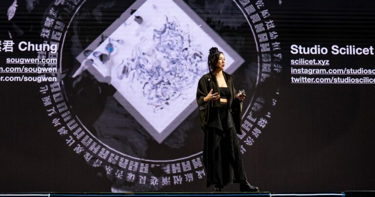Sougwen Chung, artista e pesquisadora sino-canadense, apresenta sua palestra no palco do IT Forum Praia do Forte 2025. Ela está em pé, gesticulando com as mãos enquanto fala, vestindo blazer escuro sobre blusa preta. Ao fundo, telas exibem padrões visuais em tons de azul que remetem a ondas ou fluxos digitais, criando uma atmosfera tecnológica que complementa sua apresentação sobre arte e inteligência artificial.