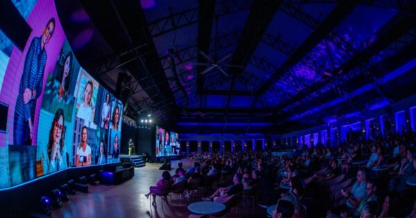 A imagem retrata um amplo auditório repleto de pessoas sentadas, todas voltadas para um palco onde ocorre uma apresentação. No palco, há várias telas grandes que exibem imagens de diferentes indivíduos. A iluminação do ambiente é composta por luzes em tons de azul e roxo, conferindo uma atmosfera moderna e tecnológica. O teto do auditório é alto e apresenta uma estrutura metálica visível. (IT Forum Praia do Forte)