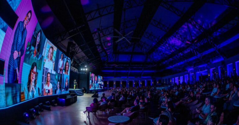 A imagem retrata um amplo auditório repleto de pessoas sentadas, todas voltadas para um palco onde ocorre uma apresentação. No palco, há várias telas grandes que exibem imagens de diferentes indivíduos. A iluminação do ambiente é composta por luzes em tons de azul e roxo, conferindo uma atmosfera moderna e tecnológica. O teto do auditório é alto e apresenta uma estrutura metálica visível. (IT Forum Praia do Forte)