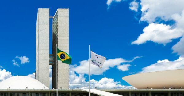 A imagem retrata o Congresso Nacional do Brasil, situado em Brasília. No centro, destacam-se duas torres retangulares e altas, que compõem os edifícios principais. À esquerda das torres, vê-se uma cúpula branca, representando a Câmara dos Deputados, enquanto à direita há uma estrutura em forma de prato invertido, que corresponde ao Senado Federal. Em primeiro plano, estão duas bandeiras hasteadas: a bandeira do Brasil à esquerda e a bandeira do Mercosul à direita. O céu ao fundo está azul com nuvens brancas esparsas, compondo um cenário claro e iluminado. (governo e ciber)